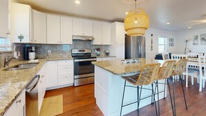 A modern kitchen with white cabinets, granite countertops, a central island with bar stools, stainless steel appliances, and a woven pendant light. - In Town, Ample Parking, Swing set for Kids, Pet Friendly (Port Aransas)