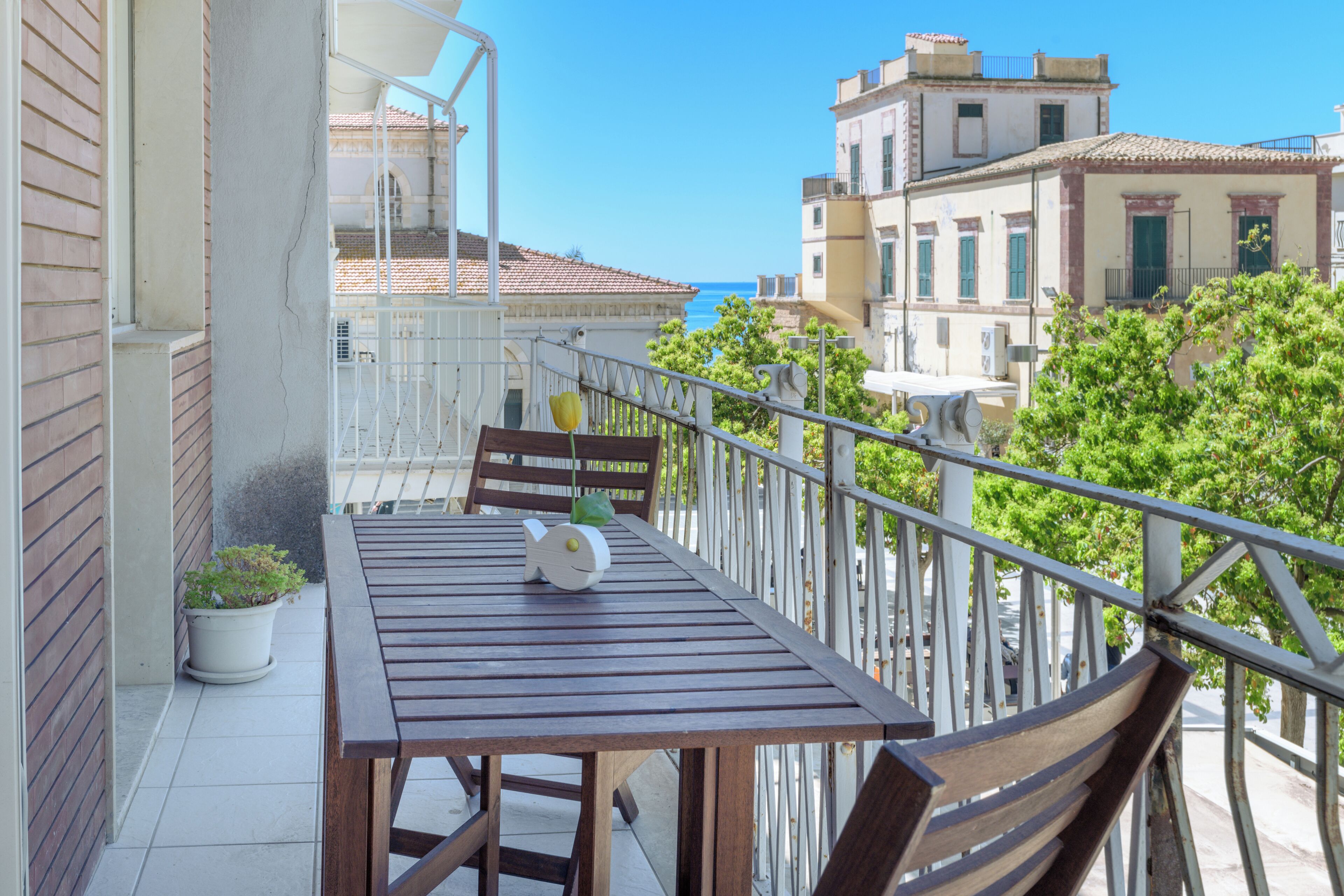 A balcony with a wooden table and chairs, a potted plant, and a view of buildings and trees.