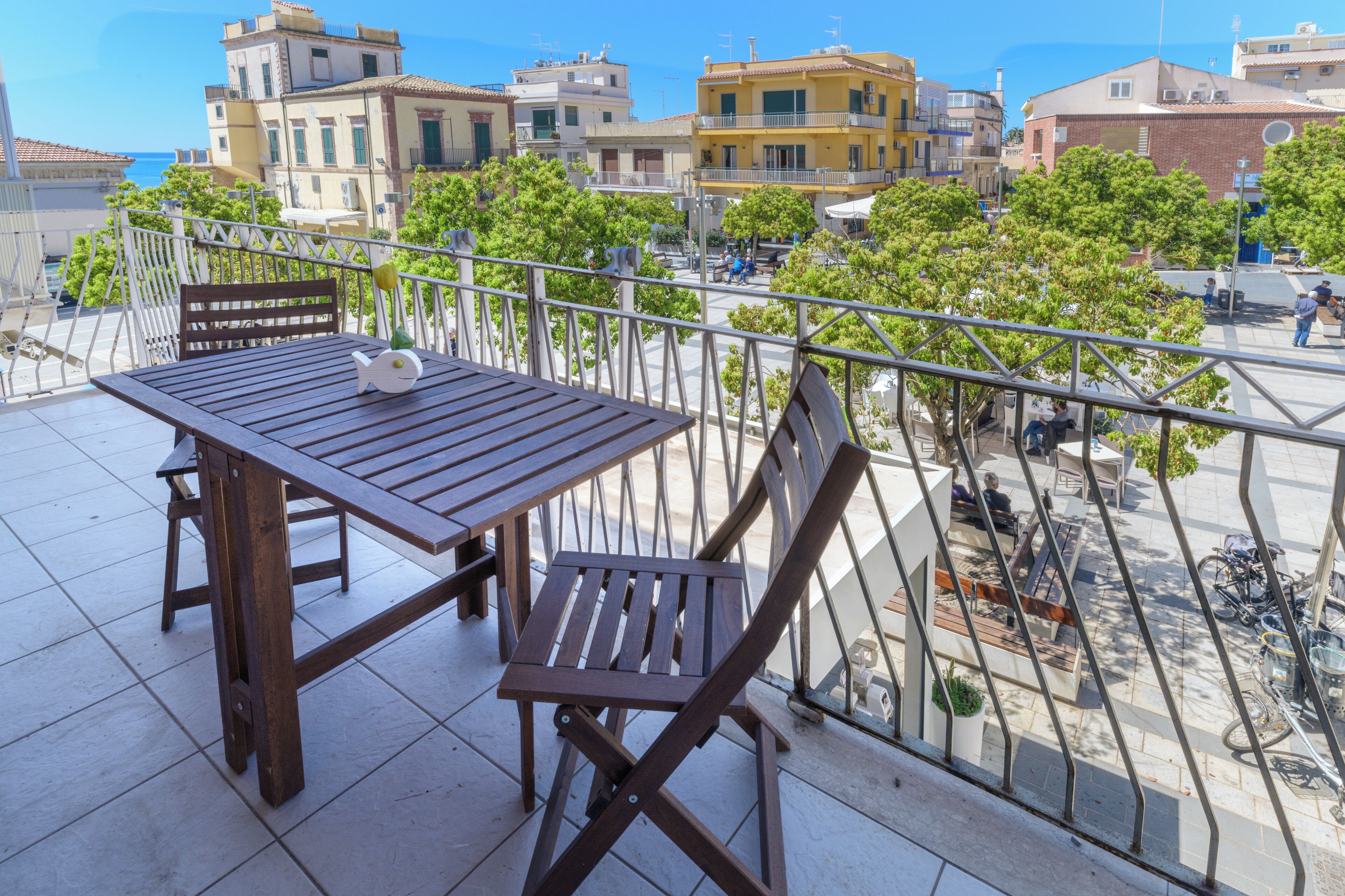 A balcony with a wooden table and chairs, overlooking a cityscape with buildings and trees.