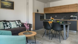 Een moderne woonkamer met een bank, salontafel en een eethoek met stoelen. In de achtergrond is een keuken met houten kasten en apparatuur zichtbaar.