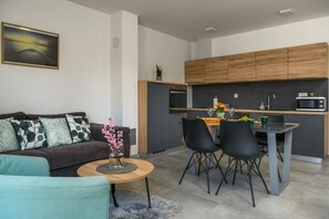 A modern living room with a sofa, coffee table, and a dining area with chairs. A kitchen with wooden cabinets and appliances is visible in the background. - NEW Villa Dora - Apartment 1 "Green" with terrace and barbecue (Pula)