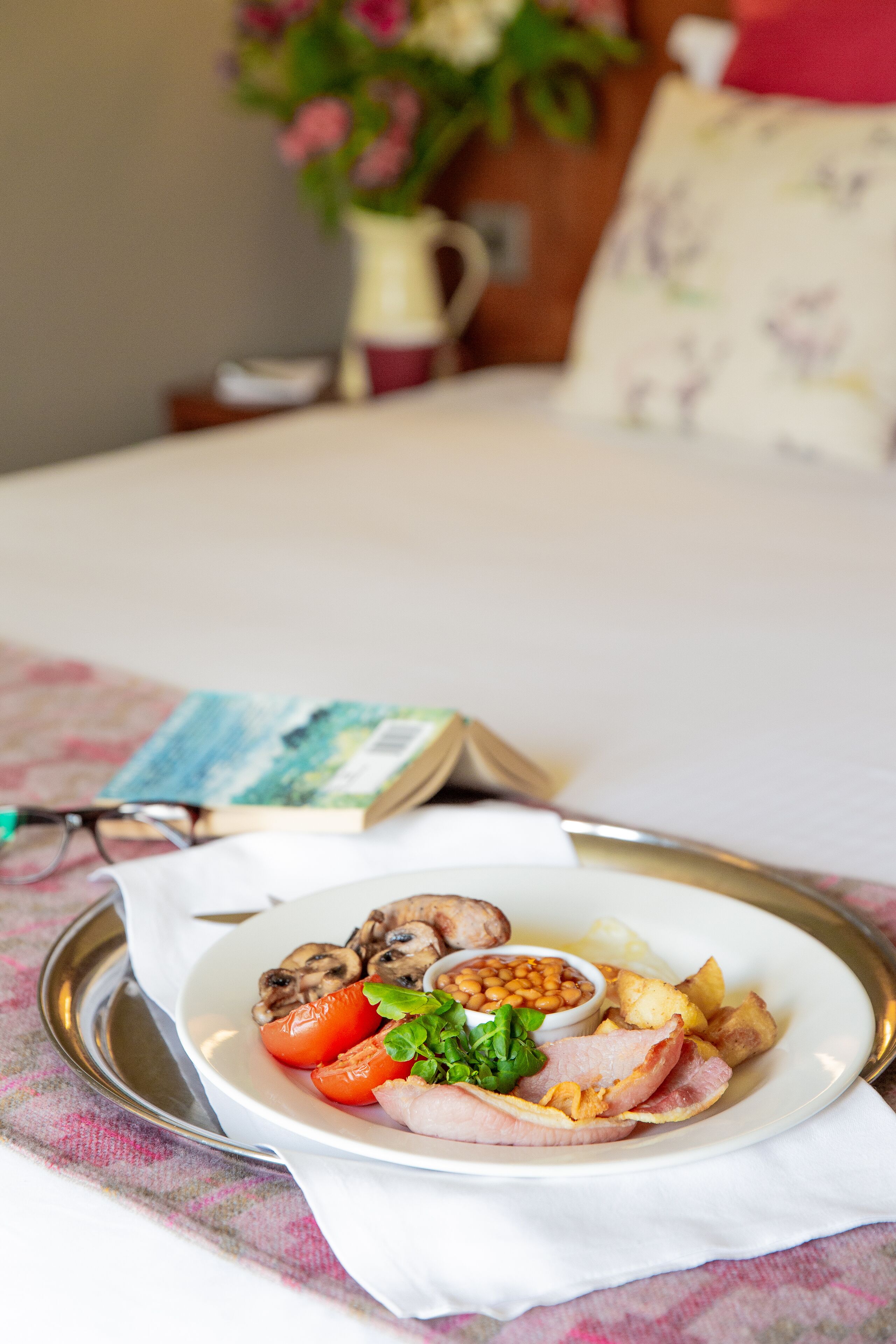 A breakfast tray with eggs, mushrooms, tomatoes, beans, and ham on a bed with a book and glasses beside it.