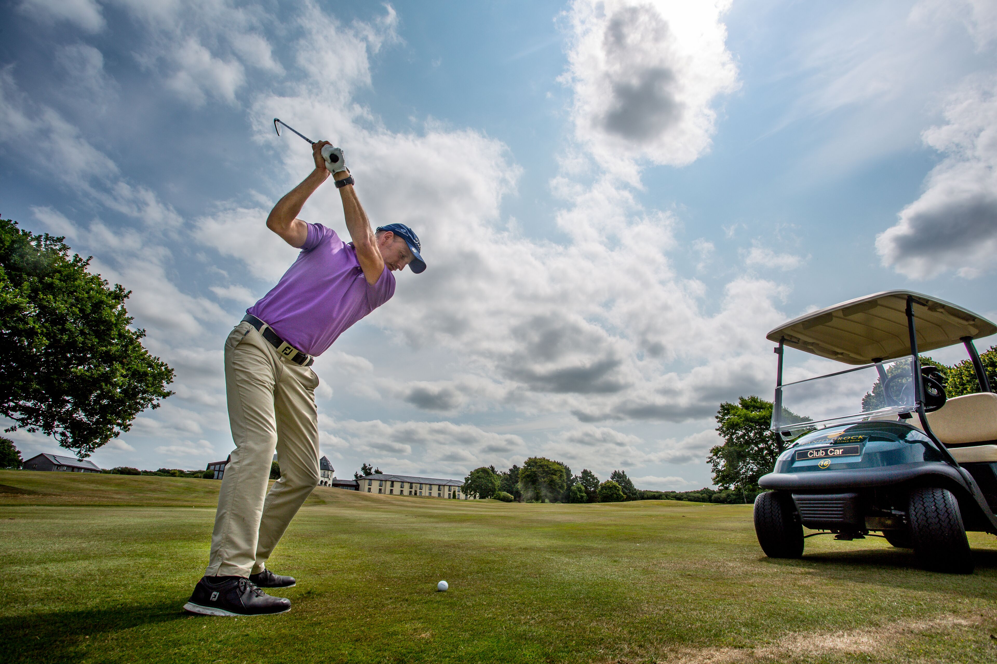 A golf course with a golf cart and a golfer in a purple shirt and khaki pants preparing to hit a golf ball.
