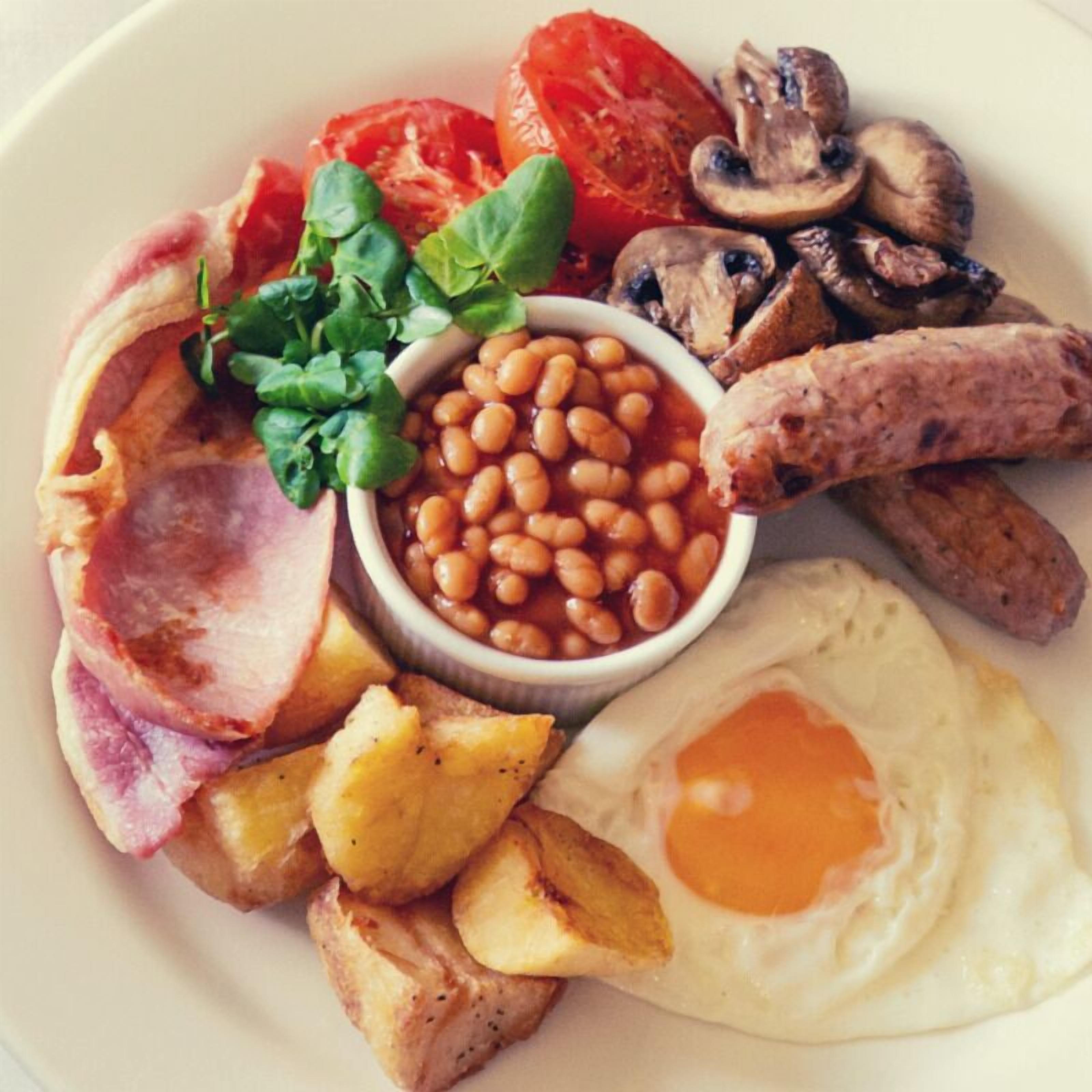 A plate of breakfast food including eggs, bacon, sausages, mushrooms, tomatoes, beans, and greens.