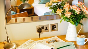 A hotel room with a wooden desk, a mirror, a lamp, a vase with flowers, a coffee cup, and a bed with pillows. - Lanhydrock Hotel and Golf Club (Bodmin)