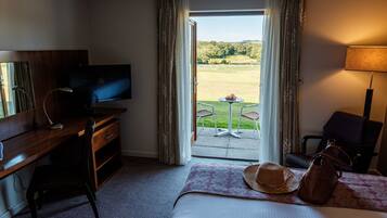 A hotel room with a bed, a desk with a TV, a laptop, a chair, a table with a lamp, and a view of a golf course through the door.