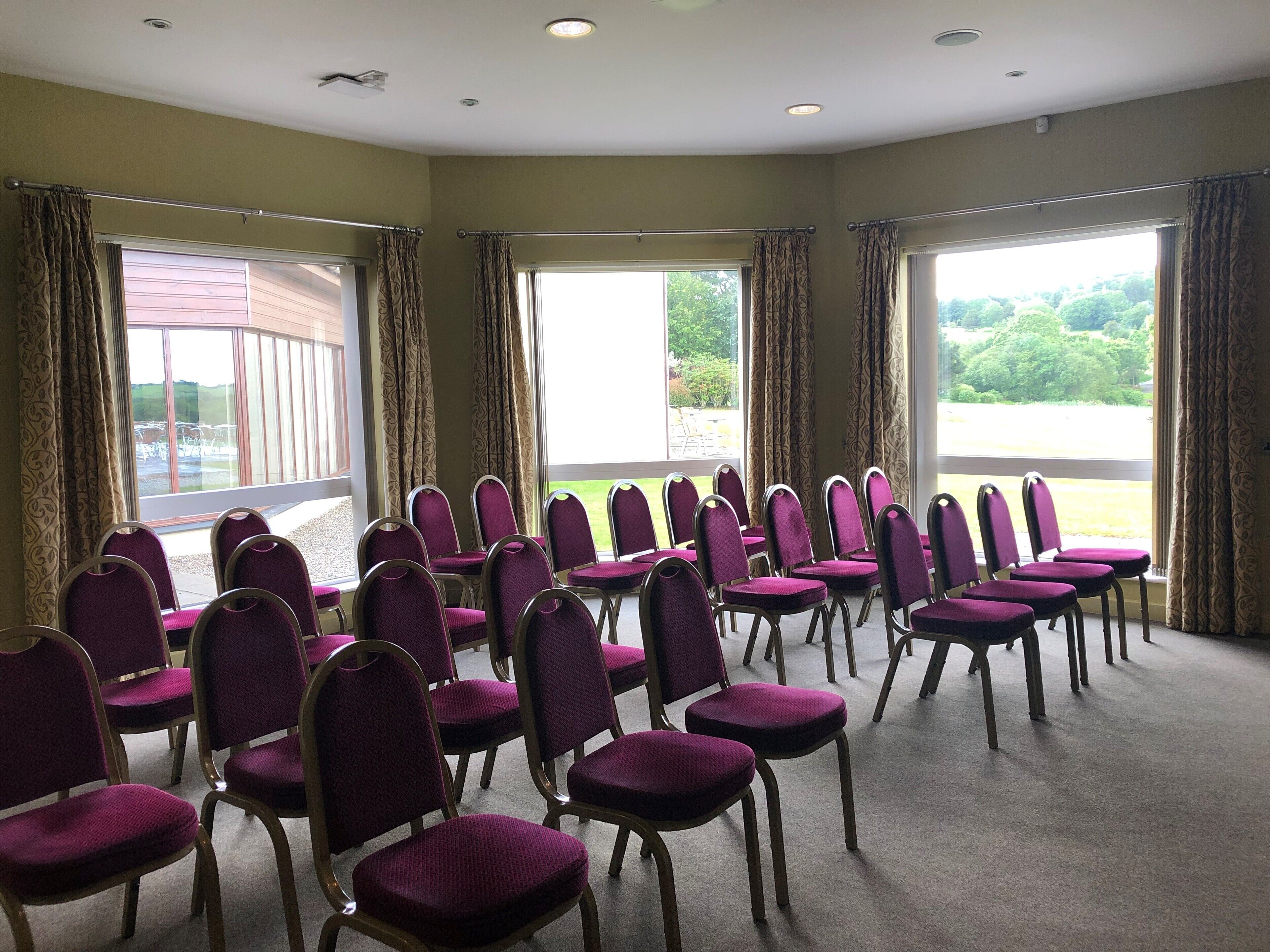 A conference room with rows of purple chairs, large windows with curtains, and a view of a building and greenery outside.