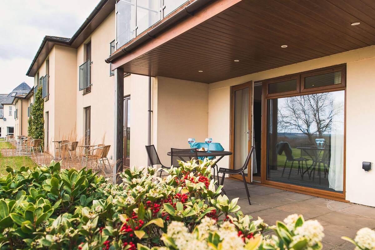 A covered patio area with outdoor furniture, a glass door leading to a garden, and a view of a residential area.