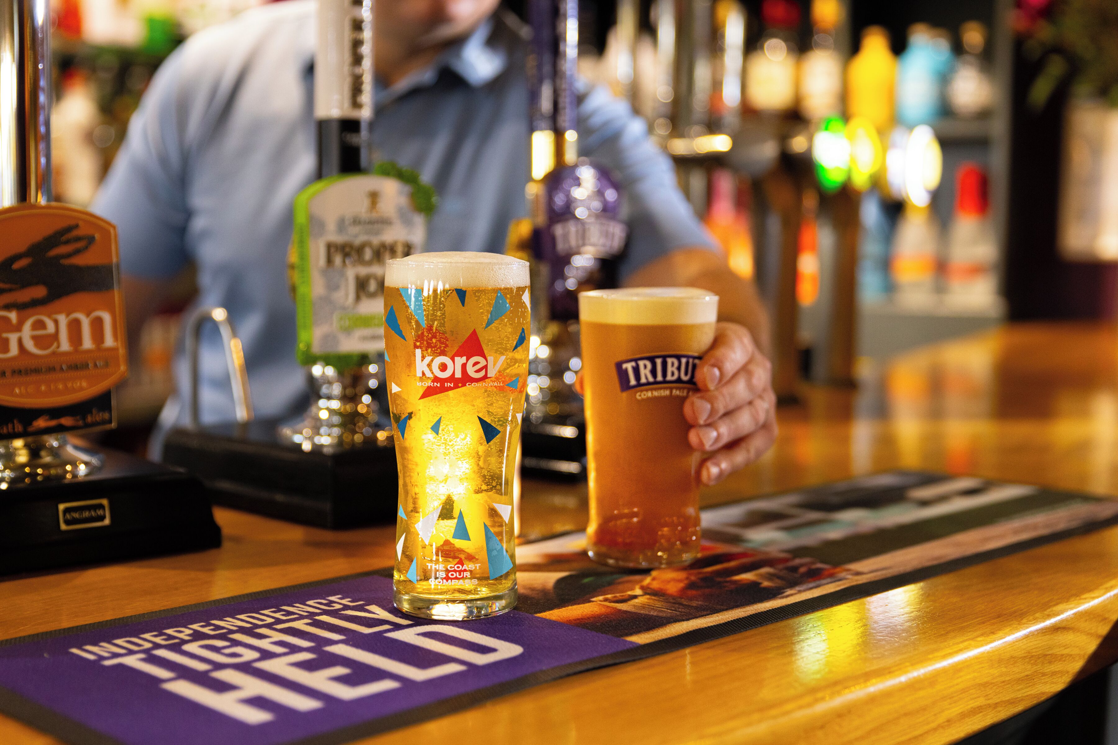 Un bar con dos vasos de cerveza, una llave de cerveza con la etiqueta 'Gem' y un posavasos con el texto 'Independence Tight Held'.