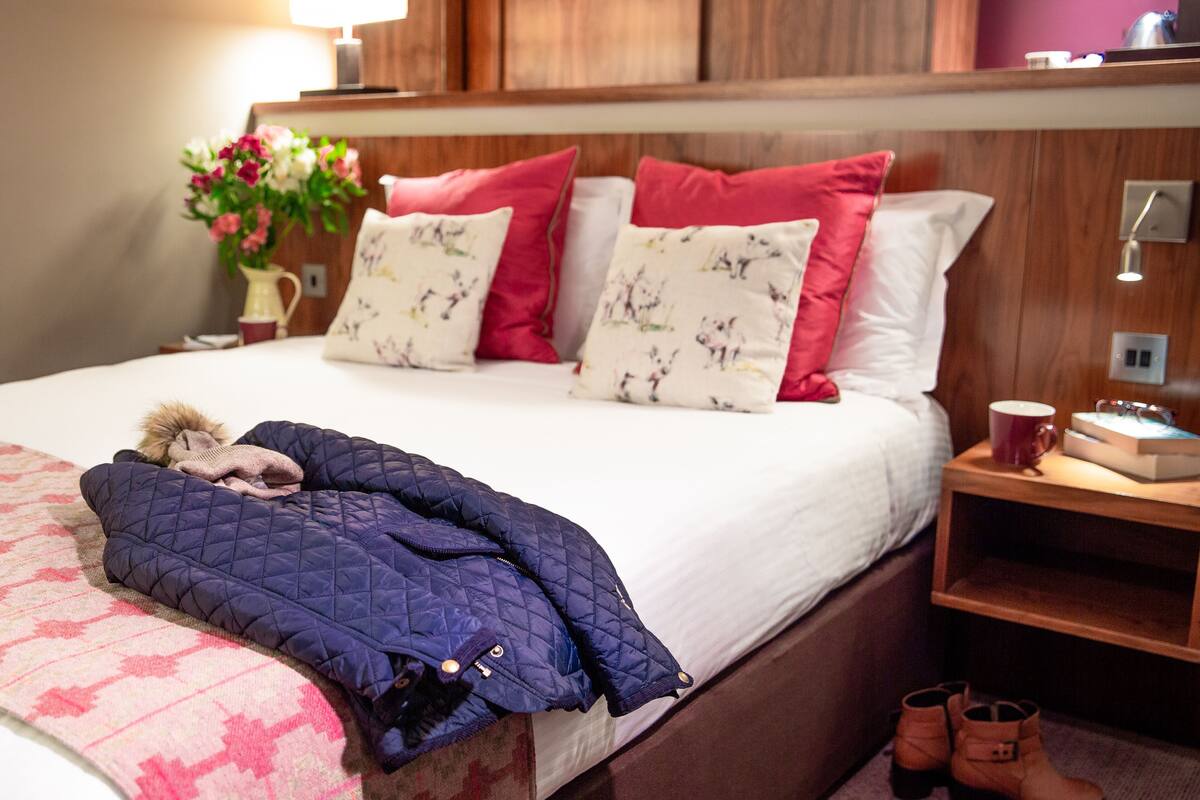 A neatly made bed with a blue quilt, red and white pillows, and a small stuffed animal. A wooden nightstand with a lamp and a red mug is beside the bed.
