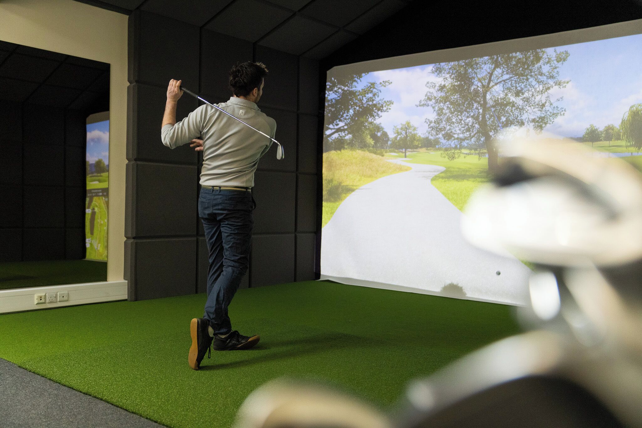 Een persoon oefent golf in een ruimte met een groot scherm waarop een golfbaan wordt getoond.
