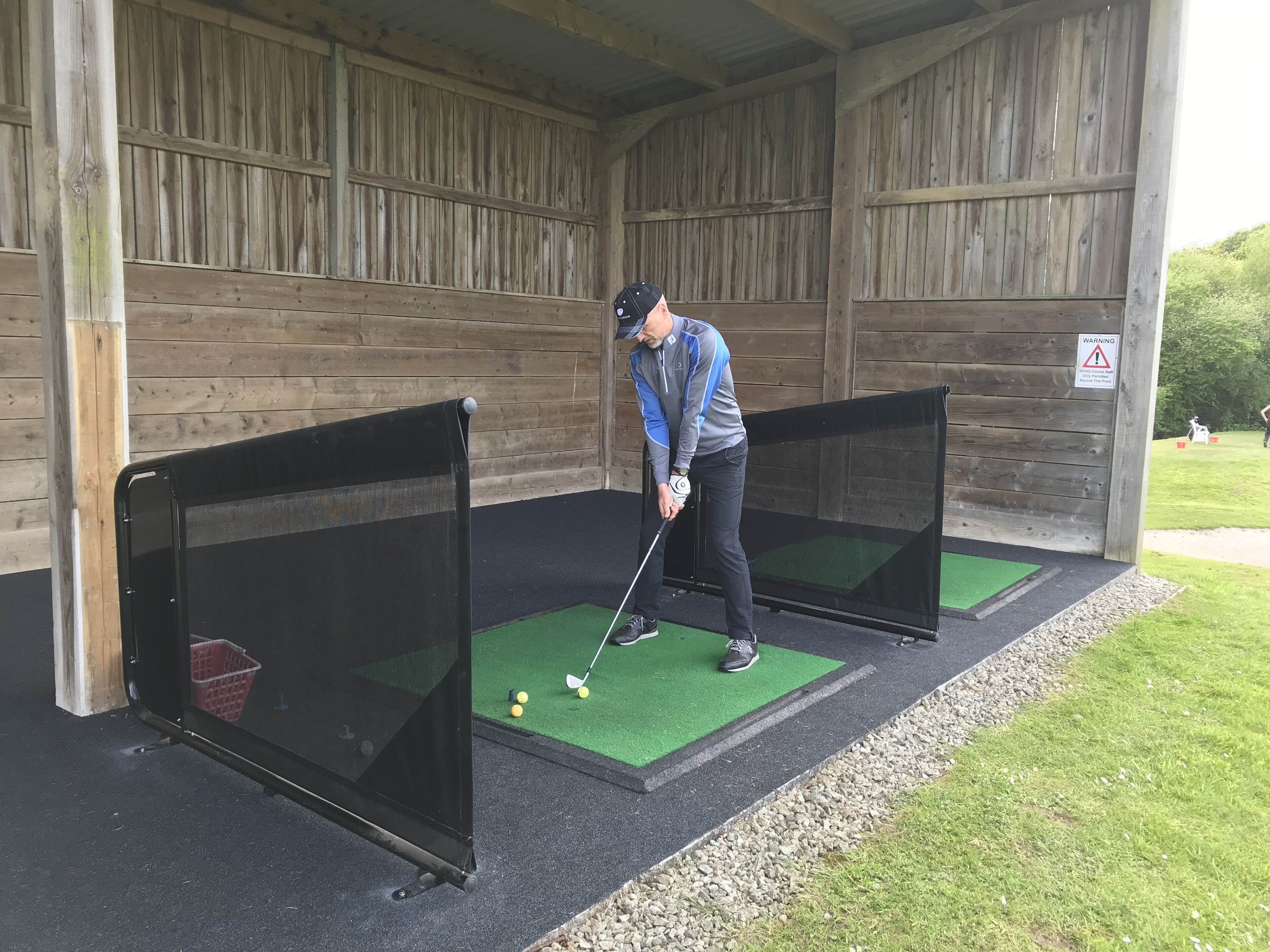 A man practicing golf in a covered area with two practice bays, a warning sign, and a grassy area outside.