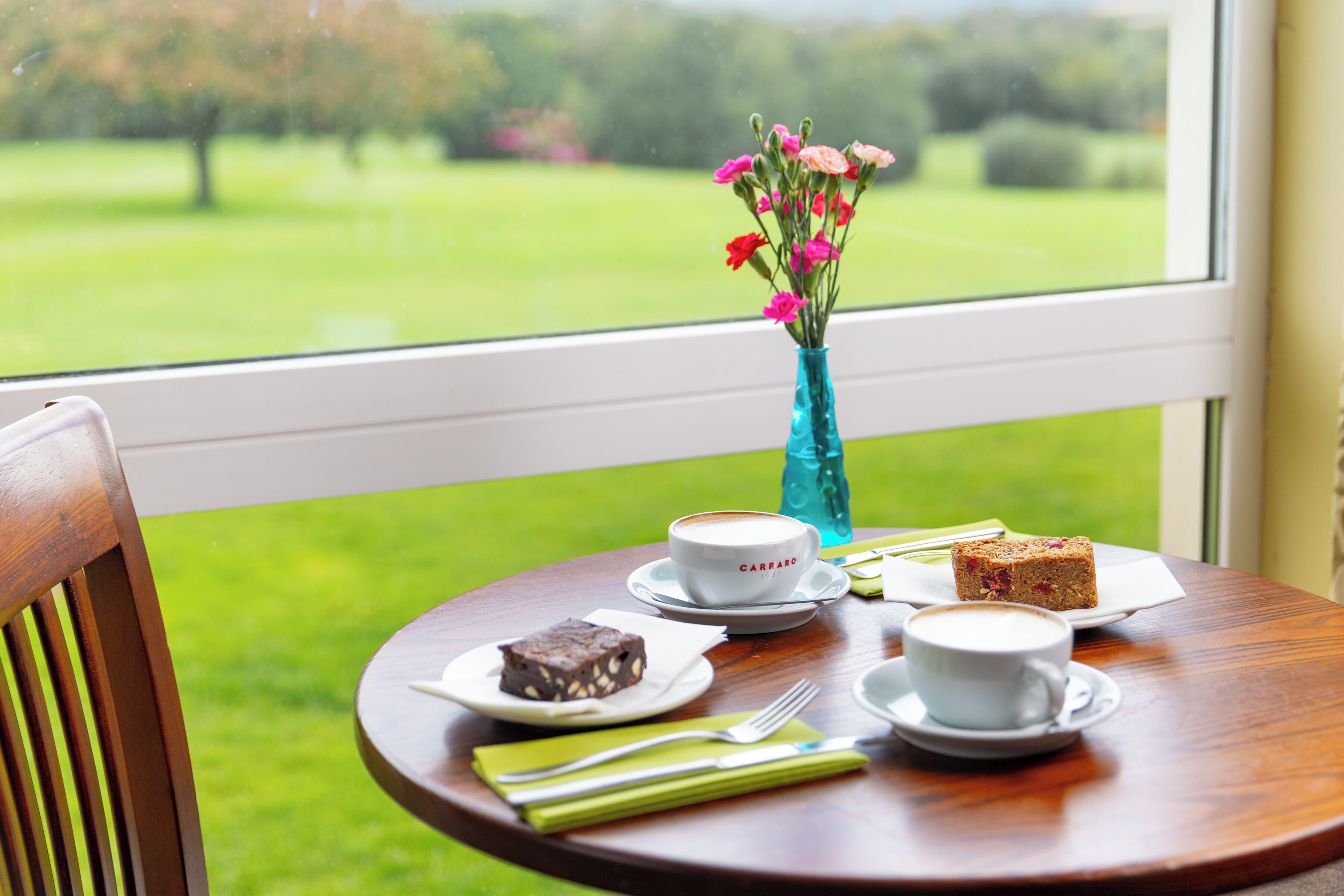Ein Tisch mit einer Kaffeetasse, einer Kuchenstück, einer Blumenvase und Blick auf eine grüne Landschaft durch ein Fenster.