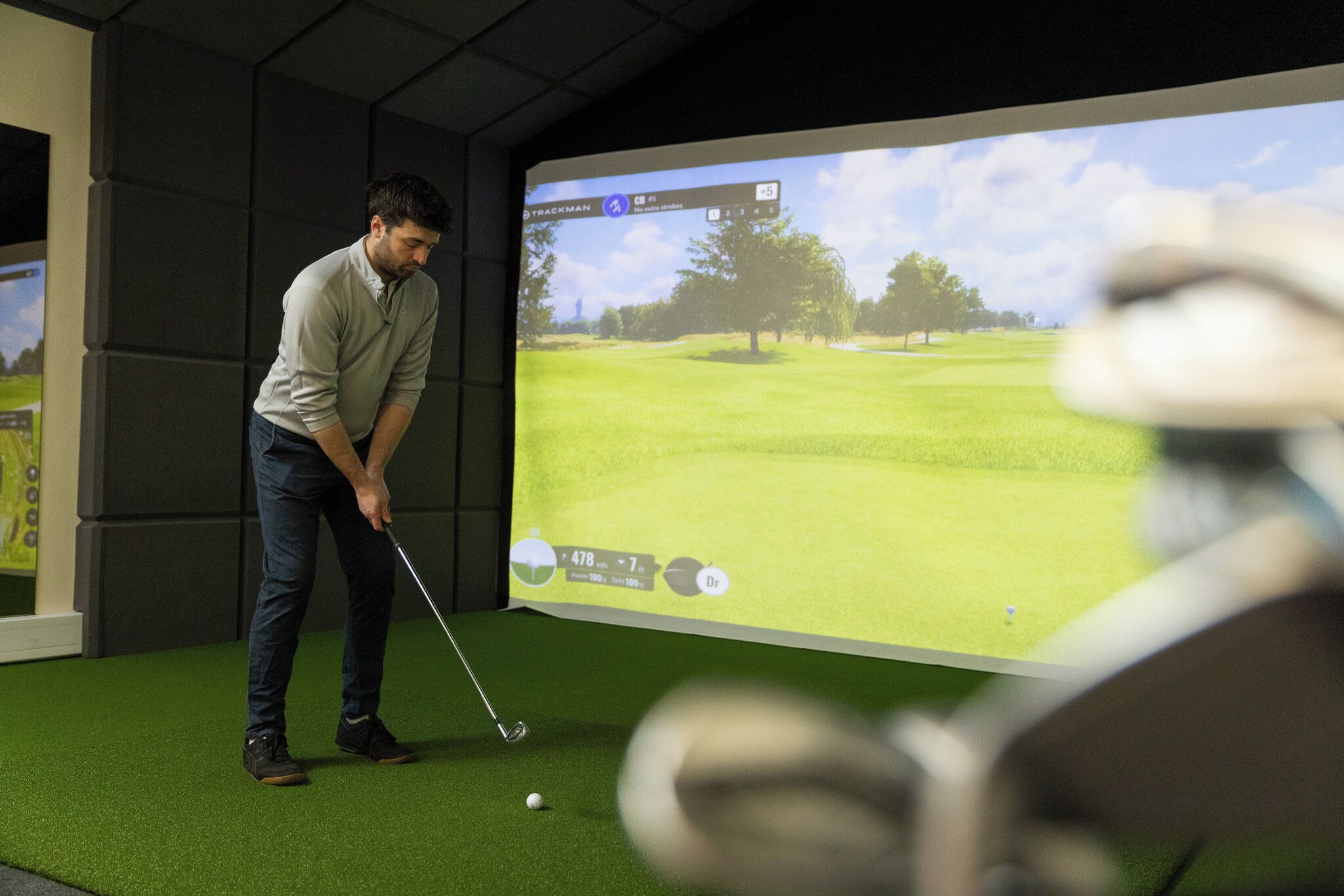 Una persona jugando golf en un espacio interior, con una pantalla grande que muestra un campo de golf.