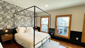 Un dormitorio con una cama con dosel, dos mesitas de noche, una mesita auxiliar, una ventana con persianas y un papel tapiz floral.