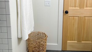Un baño con pared de azulejos blancos, una toalla blanca colgada en un perchero, una cesta de mimbre, una puerta de madera y un cartel enmarcado en la pared.
