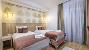 Hotellihuone, jossa on kaksi sänkyä, puinen pääty, yöpöytä ja ikkuna, jossa on läpikuultavat verhot.