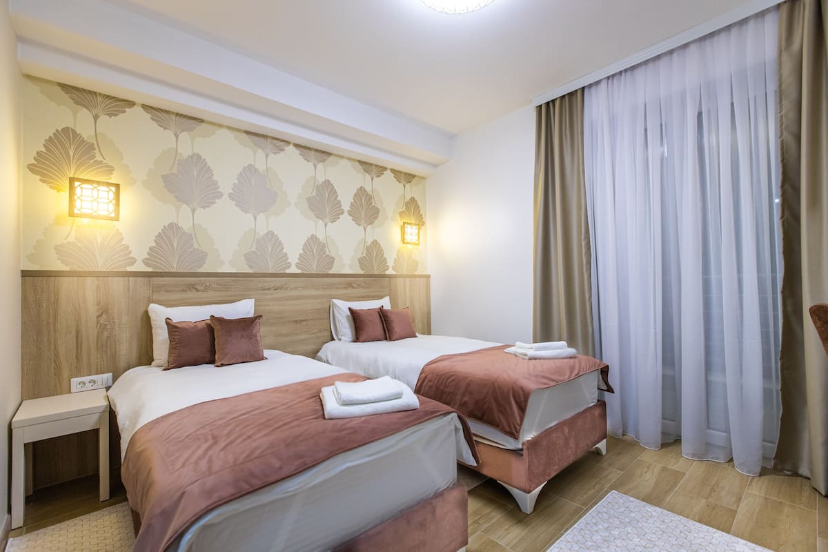 Hotellihuone, jossa on kaksi sänkyä, puinen pääty, yöpöytä ja ikkuna, jossa on läpikuultavat verhot.