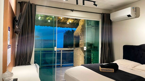 A bedroom with a bed, a small table, and a chair. There is a sliding glass door leading to an outdoor area with a view of palm trees. - The Férias - Suítes Luxo (Cabo Frio)