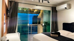 A bedroom with a bed, a small table, and a chair. There is a sliding glass door leading to an outdoor area with a view of palm trees. - The Férias - Suítes Luxo (Cabo Frio)