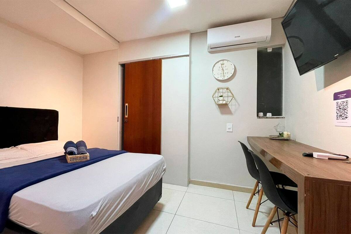 A bedroom with a bed, a desk, a chair, an air conditioner, a clock, and a wall-mounted TV.