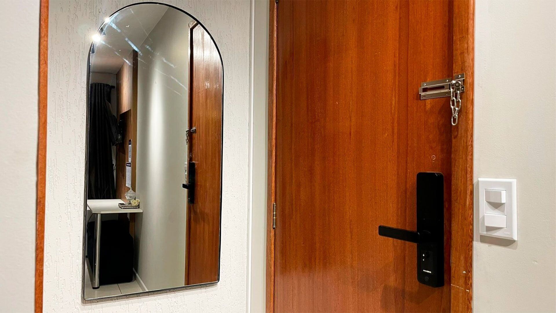 Uma porta de madeira com maçaneta preta e fechadura, uma parede branca com tomada, e um espelho refletindo parte do quarto.