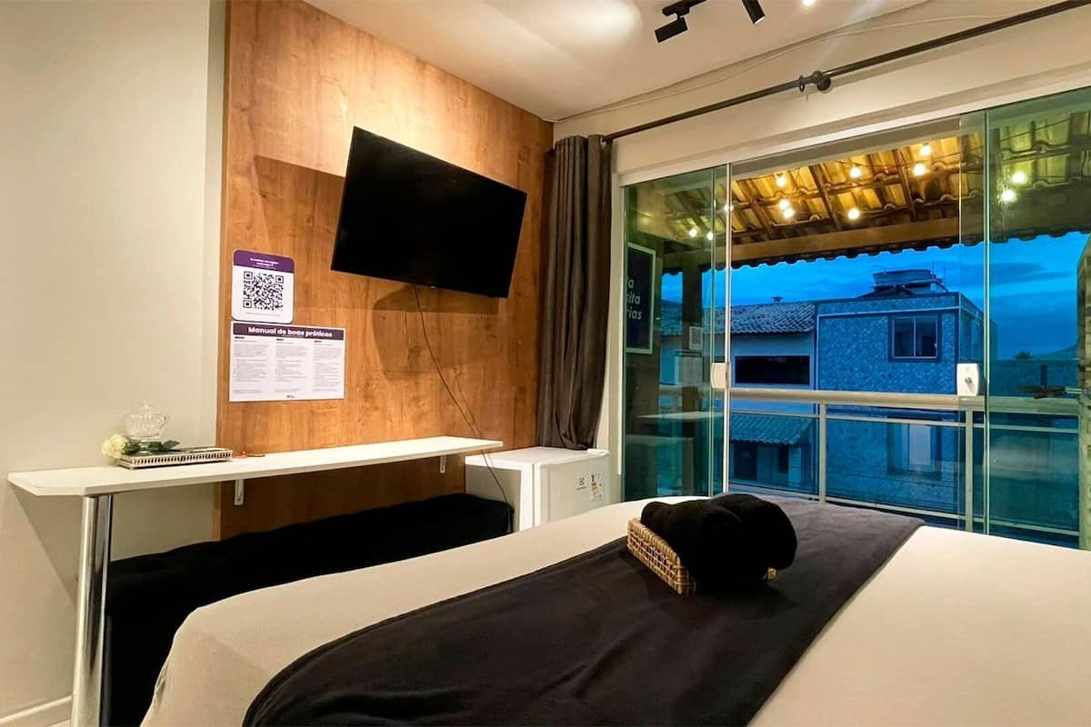 A hotel room with a bed, a desk, a flat-screen TV mounted on the wall, and a window with a view of buildings outside.