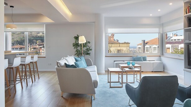 A modern living room with a sofa, chairs, a coffee table, and a view of the city through large windows. - Nautiko by FeelFree Rentals (Donostia)