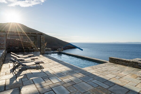 A poolside area with lounge chairs, a stone wall, and a view of the sea.