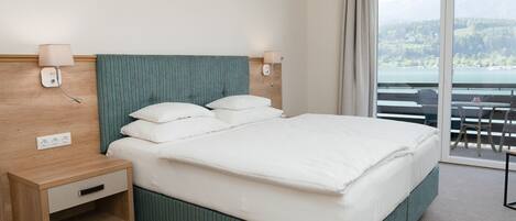 Hotelkamer met een groot bed, een houten nachtkastje, twee wandlampen en een balkon met uitzicht op een meer en bergen.