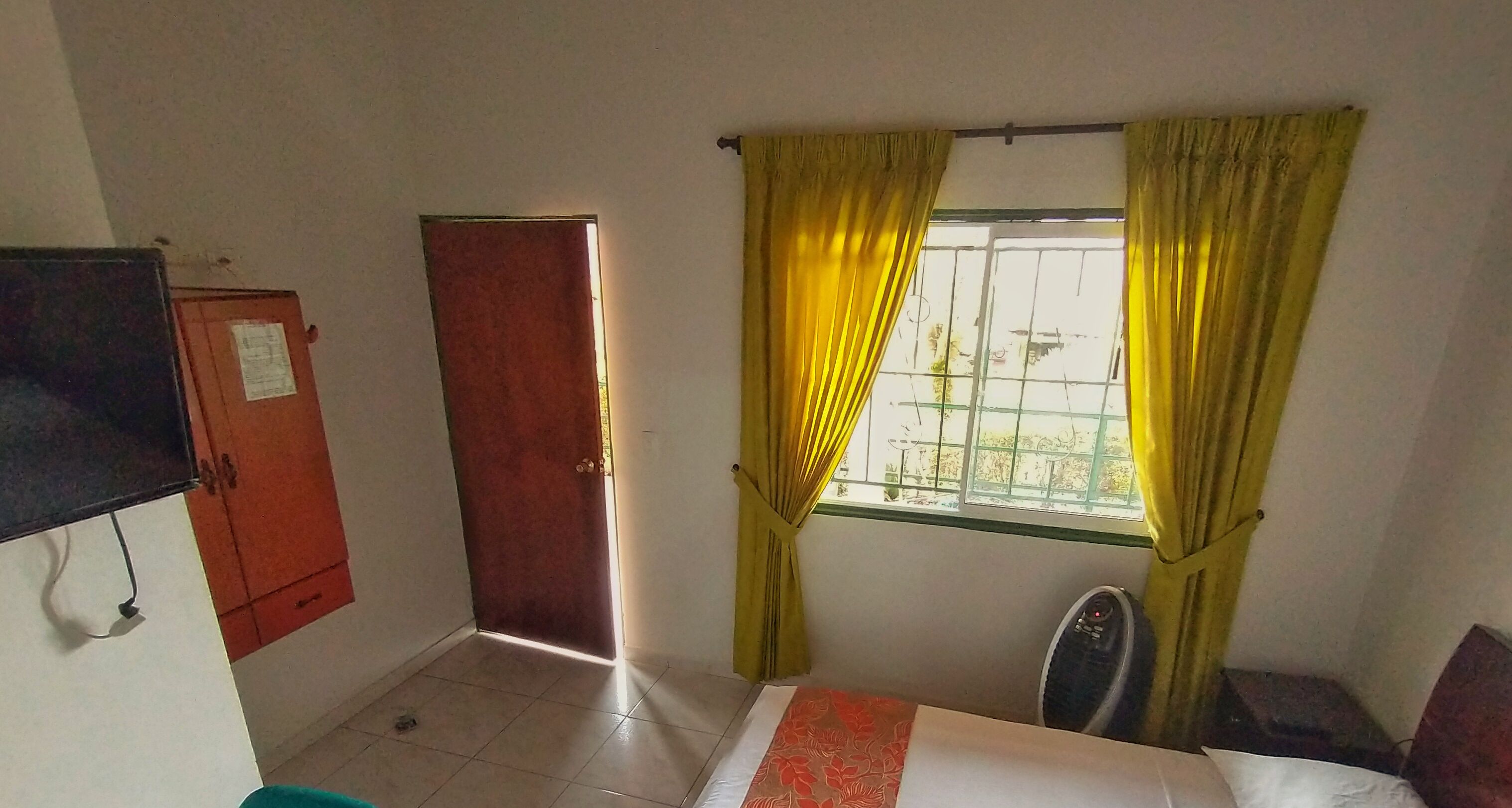 Una habitación con cama, televisión, armario, ventana con cortinas amarillas y puerta.
