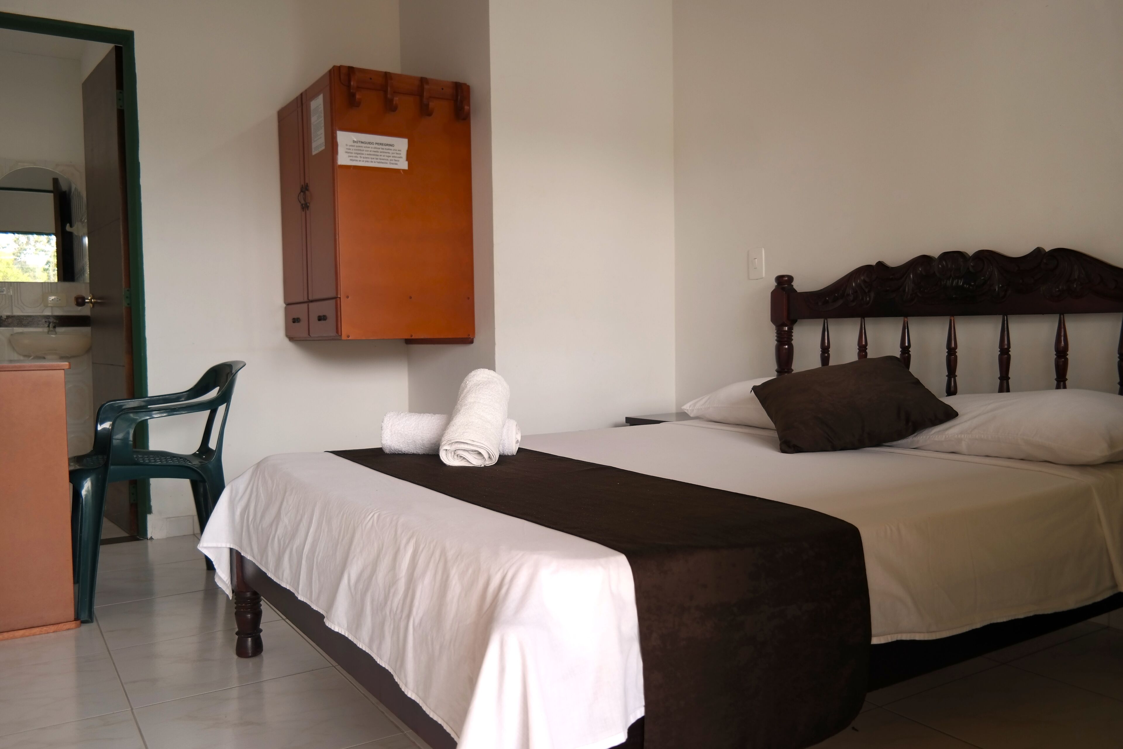 Een hotelkamer met een bed, een houten hoofdbord, een groene plastic stoel en een kastje met een bordje.