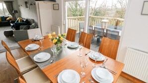 A dining area with a wooden table set for four, surrounded by chairs, with a view of the outdoors through large windows.