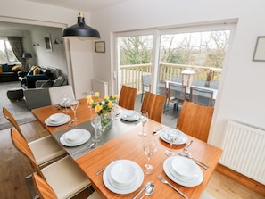 A dining area with a wooden table set for four, surrounded by chairs, with a view of the outdoors through large windows.