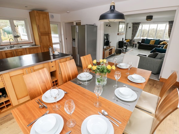 A modern kitchen with wooden cabinets, a stainless steel refrigerator, and a dining area set for six with a vase of yellow flowers.
