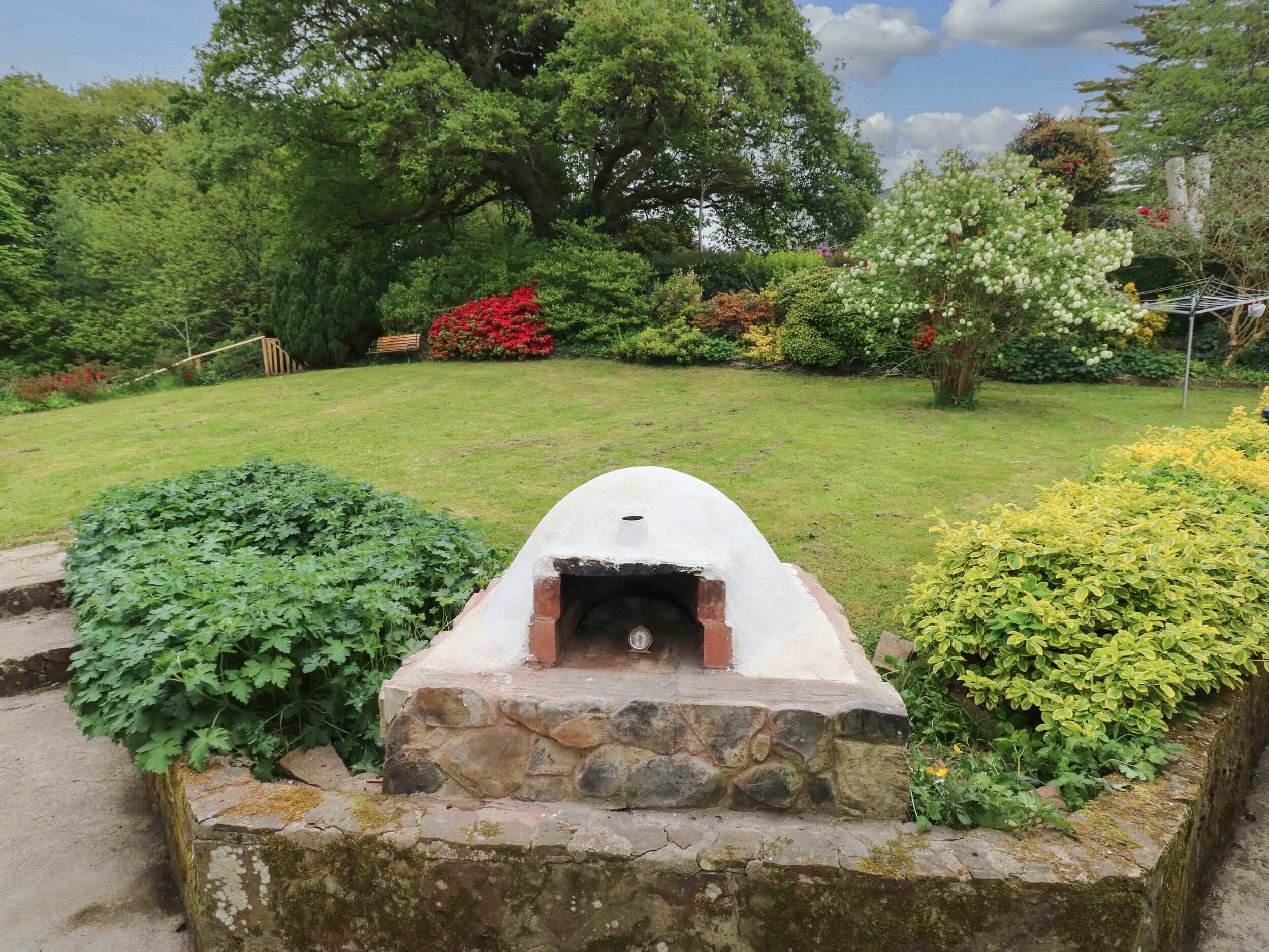 A stone-built outdoor oven in a garden with green foliage and trees in the background.
