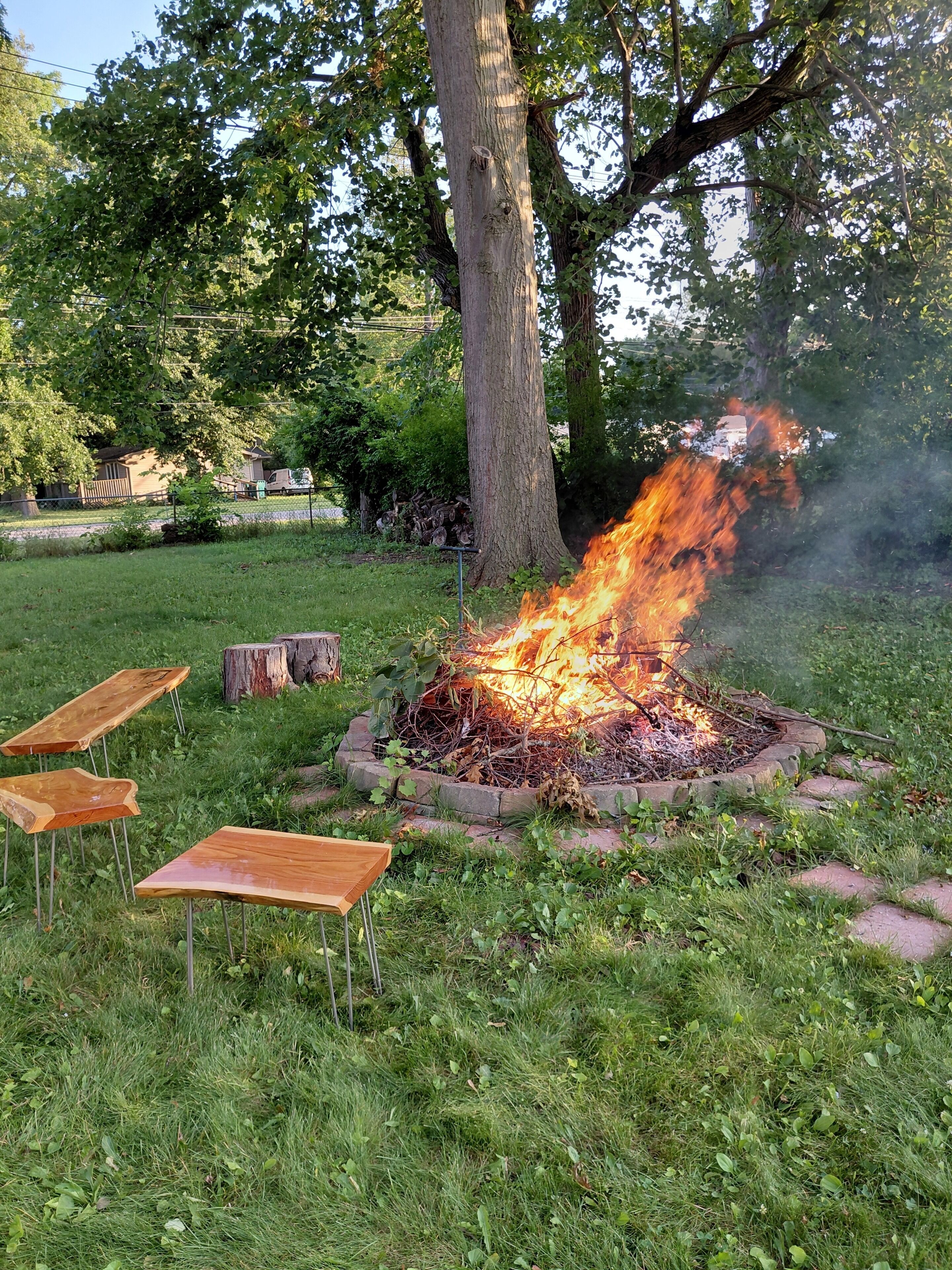 Een vuurkuil met brandende houtblokken, omringd door gras en bomen, met houten banken in de buurt.