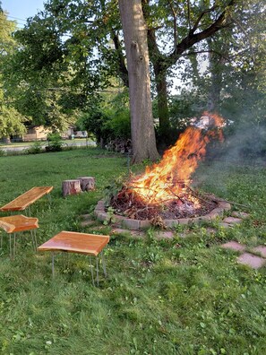 A fire pit with burning logs surrounded by grass and trees, with wooden benches nearby.