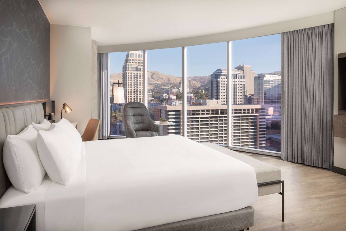 Une chambre d’hôtel moderne dotée d’un grand lit, d’une chaise, d’un bureau et d’une grande fenêtre donnant sur la ville.