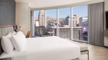 A modern hotel room with a large bed, a chair, a desk, and a large window offering a city view.