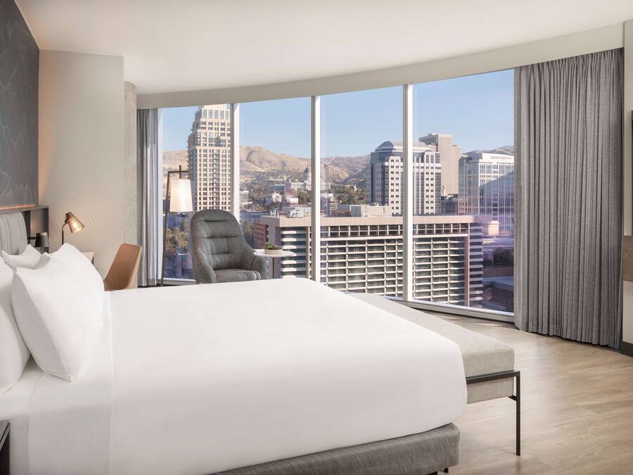 A modern hotel room with a large bed, a chair, a desk, and a large window offering a city view.