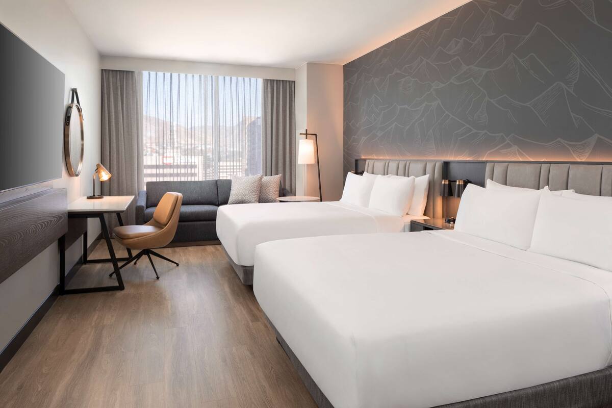 Une chambre d’hôtel avec deux lits, un bureau, une chaise, une lampe et une vue sur la ville grâce à de grandes fenêtres.