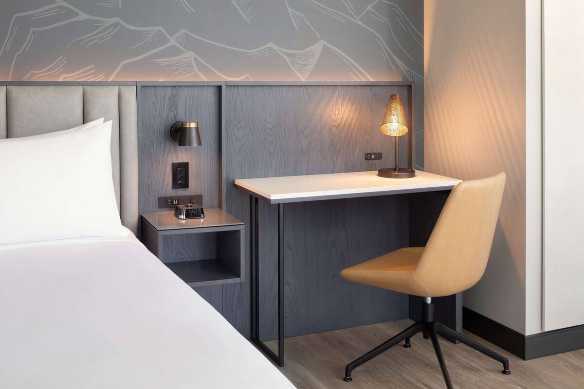 Une chambre d’hôtel comprenant un lit, un bureau avec une chaise, une table de chevet et une lampe fixée au mur.