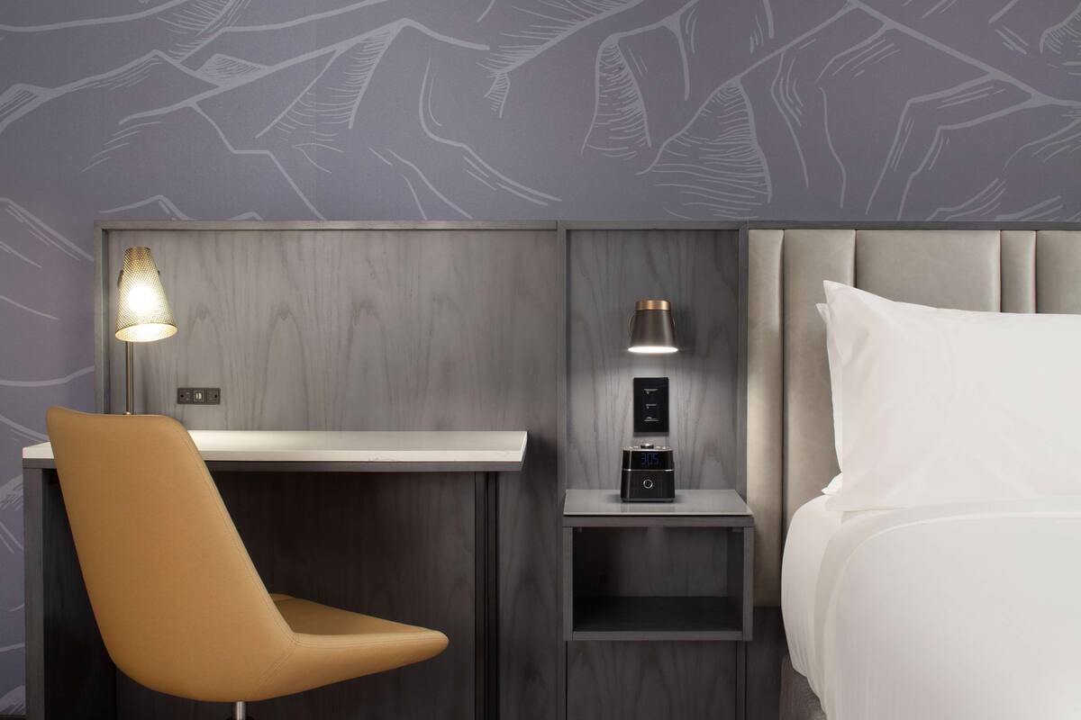 Une chambre d’hôtel moderne avec une tête de lit en bois, un bureau avec une lampe, une chaise et un lit recouvert de draps blancs.