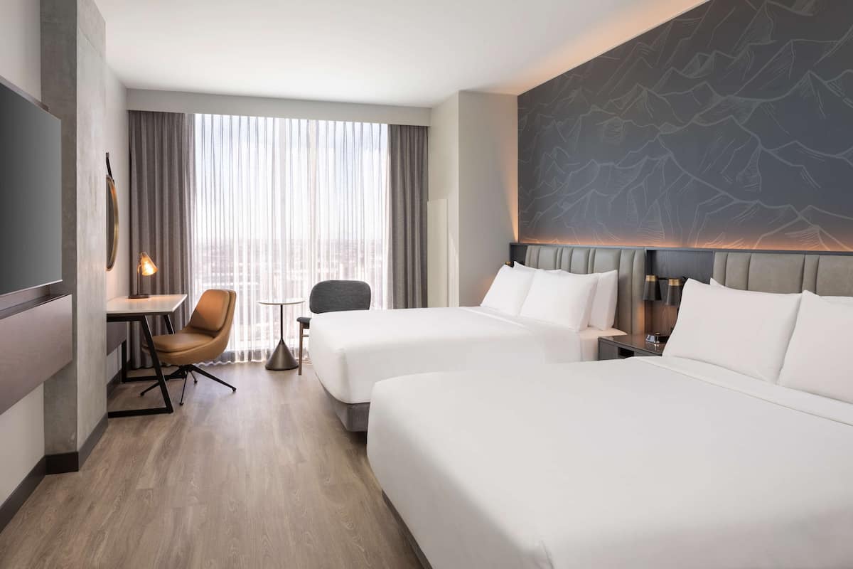 Une chambre d’hôtel avec deux lits, un bureau, une chaise, une lampe et une grande fenêtre avec des rideaux.