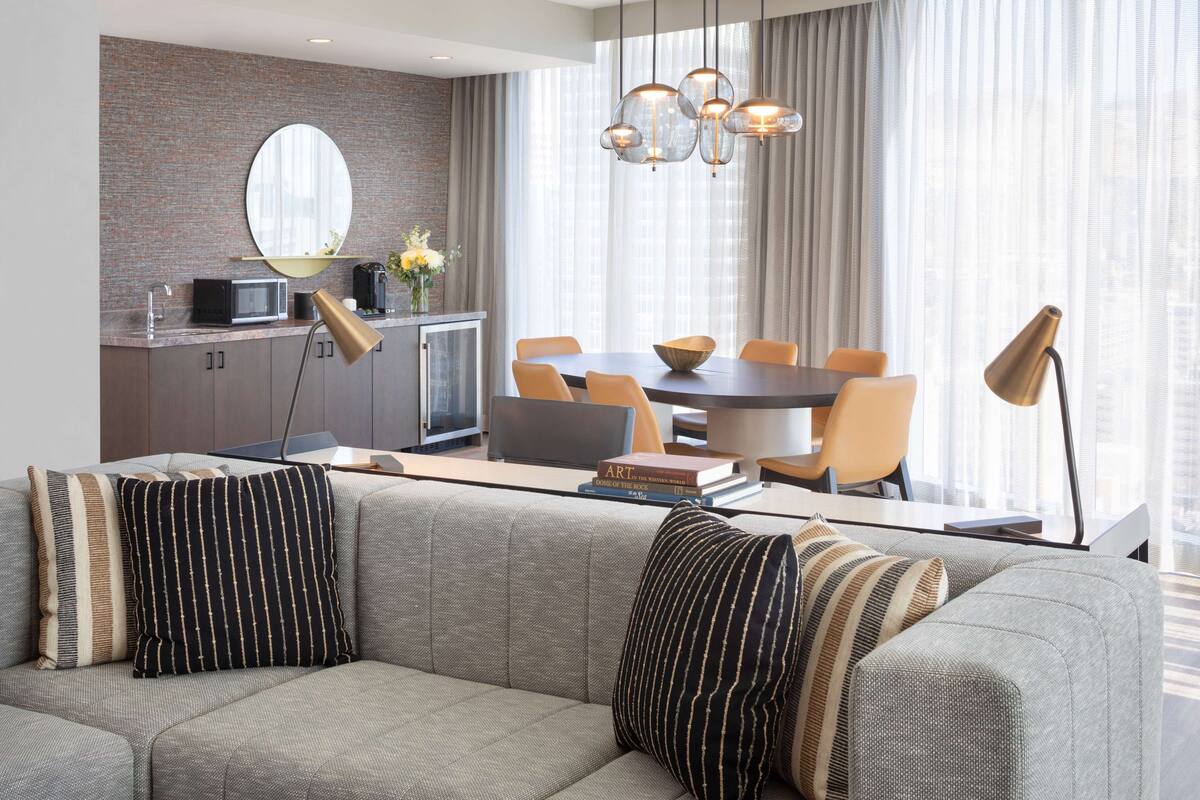 Un salon moderne avec un canapé gris, des coussins à rayures, une table à manger avec des chaises, un coin cuisine équipé d’un four à micro-ondes et une grande fenêtre avec des rideaux.
