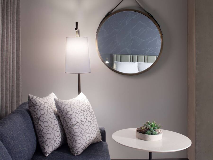Een moderne woonkamer met een grijze bank, gestreepte kussens, een ronde spiegel, een vloerlamp en een klein tafeltje met een vetplant.