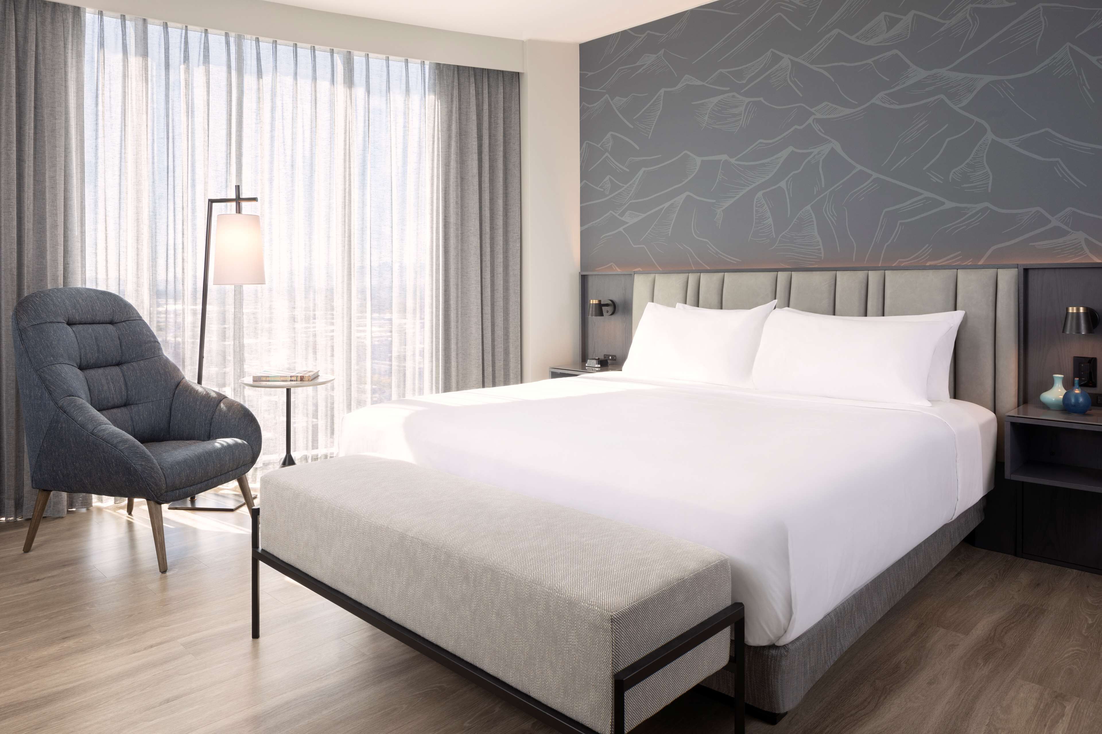 A modern hotel room with a large bed, a grey upholstered armchair, a bedside table with a lamp, and a city view through sheer curtains.