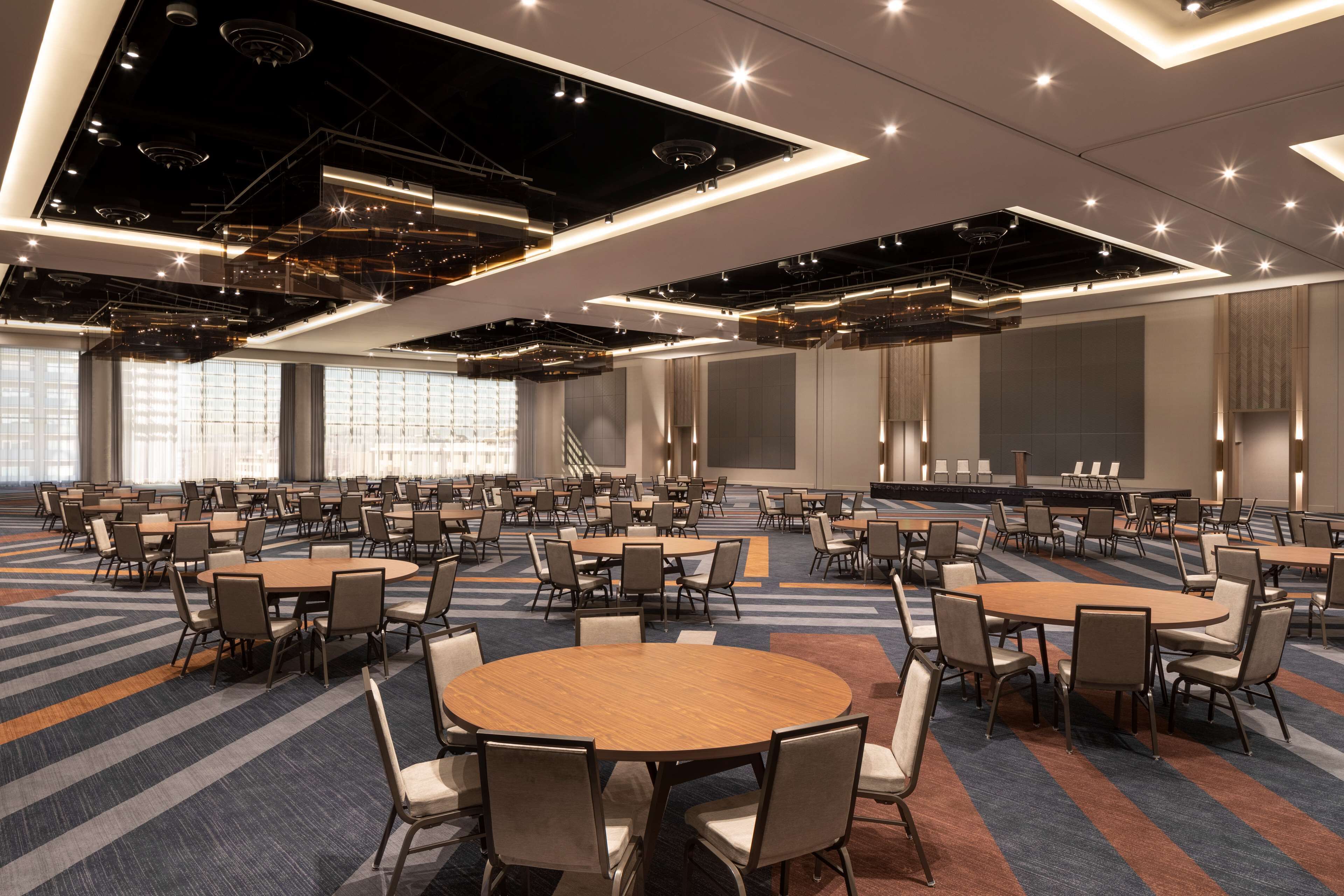 A large conference room with multiple tables and chairs, high ceilings with exposed ductwork, and large windows providing natural light.