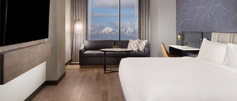 Ein Hotelzimmer mit einem großen Bett, einer Sitzecke, einem kleinen Tisch, einem Fernseher und Blick auf Berge durch das Fenster.